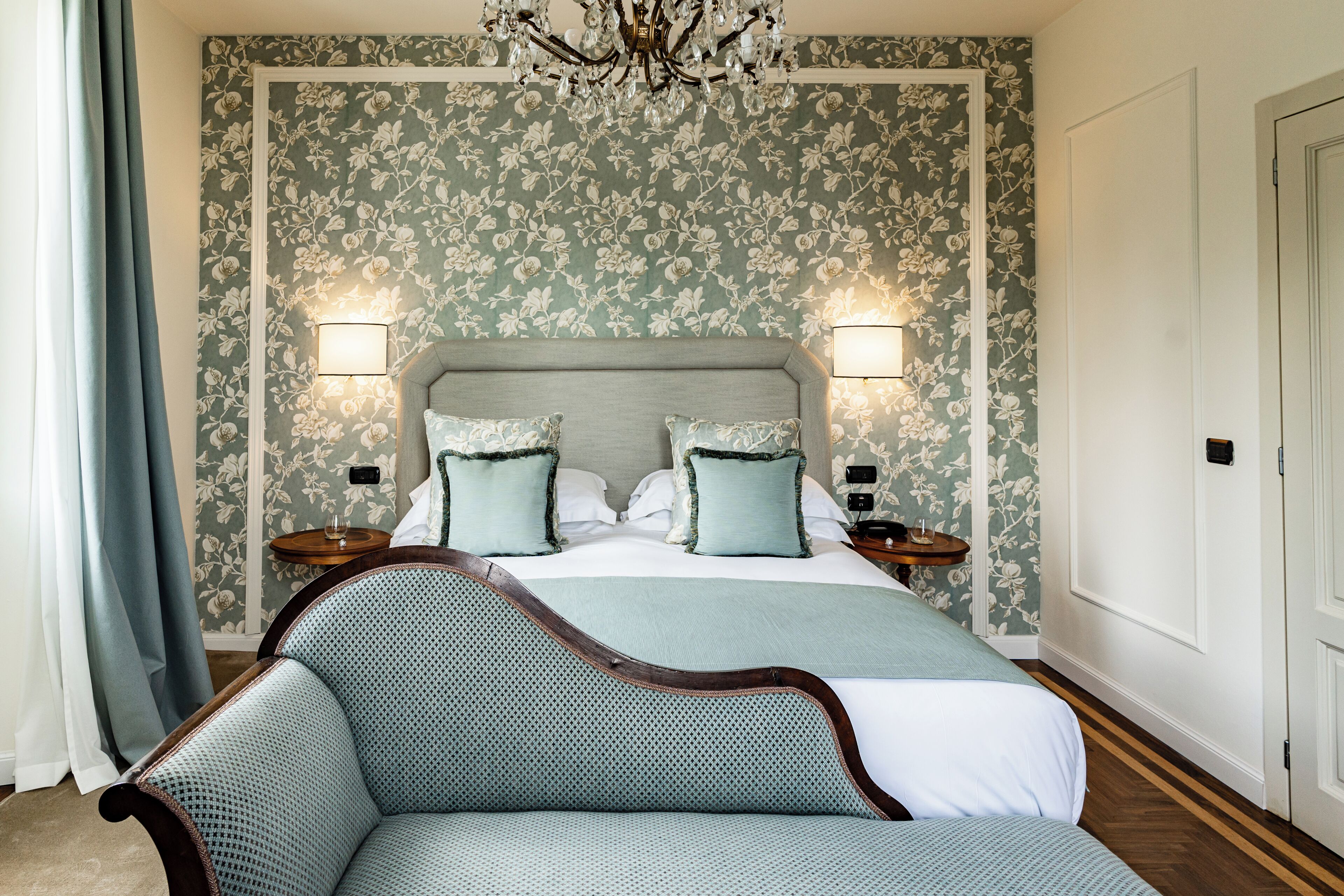 A bedroom with a large bed, two bedside tables, a chandelier, and floral wallpaper.