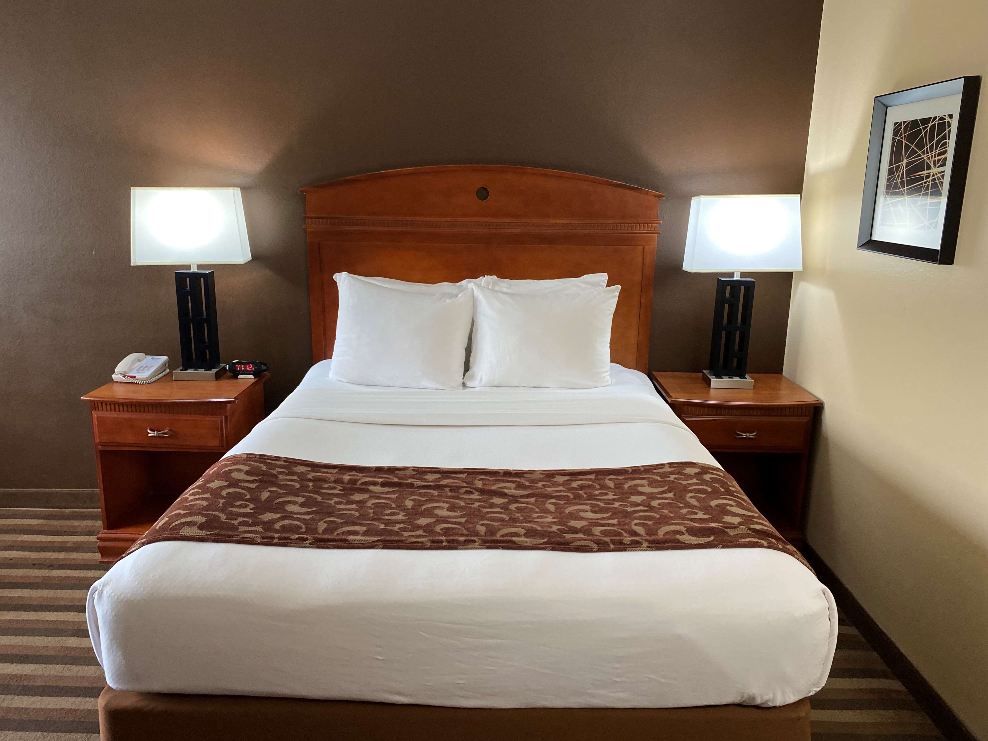 A hotel room with a wooden headboard, two bedside tables with lamps, a telephone, and a framed picture on the wall.