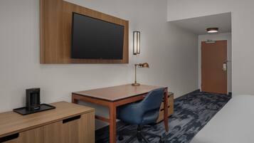 A hotel room with a wooden desk, a blue office chair, a flat-screen TV mounted on the wall, a coffee maker on the desk, and a bed with a white comforter.