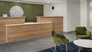 Un área de recepción con un mostrador de madera, dos sillas verdes, una mesa redonda y un elemento decorativo en la pared.