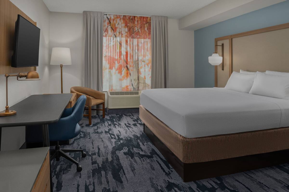 Habitación de hotel con cama, escritorio, silla, televisión y ventana con cortinas.