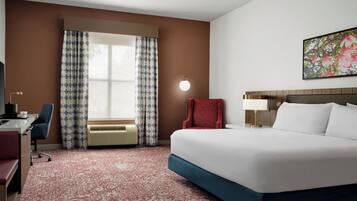 A hotel room with a large bed, a desk with a chair, a red chair, a wall-mounted lamp, and a window with patterned curtains.
