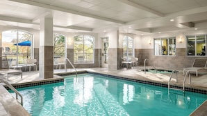 A swimming pool with clear water, surrounded by lounge chairs and tables, and large windows offering views of the outdoors. - Hilton Garden Inn Jackson/Madison (Madison)