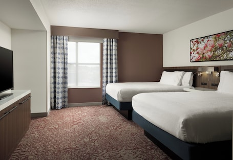 A hotel room with two beds, a flat-screen TV, a large window with patterned curtains, and a floral artwork on the wall.