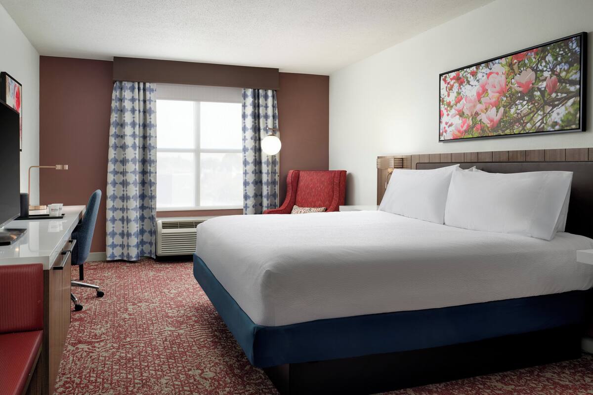 A hotel room with a large bed, a desk, a chair, a red armchair, a window with curtains, and a wall-mounted picture.