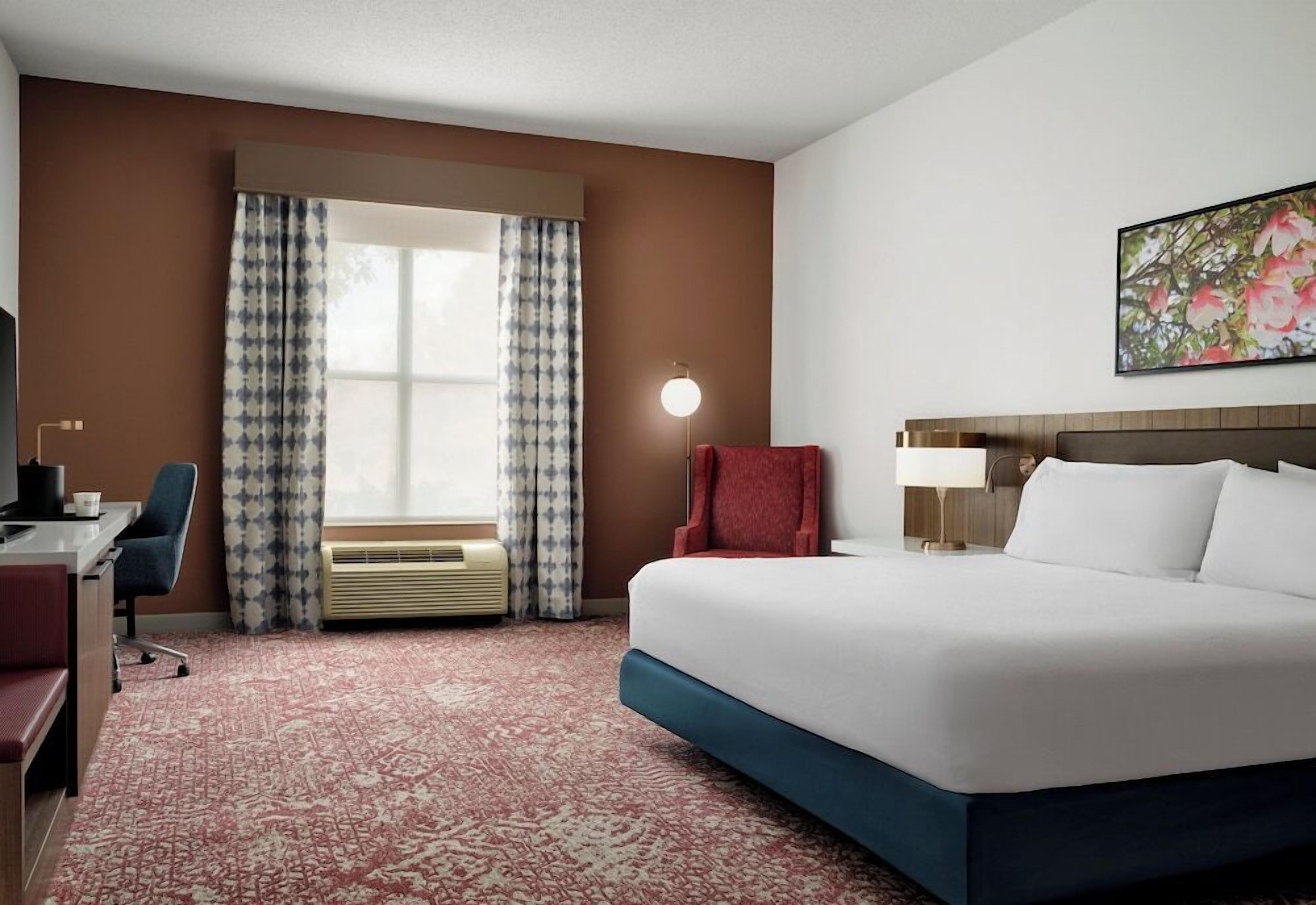 A hotel room with a large bed, a desk, a chair, a lamp, and a window with curtains.
