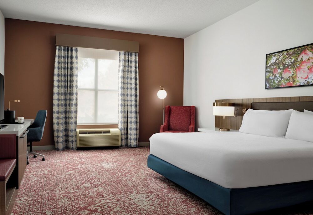 A hotel room with a large bed, a desk, a chair, a lamp, and a window with curtains.