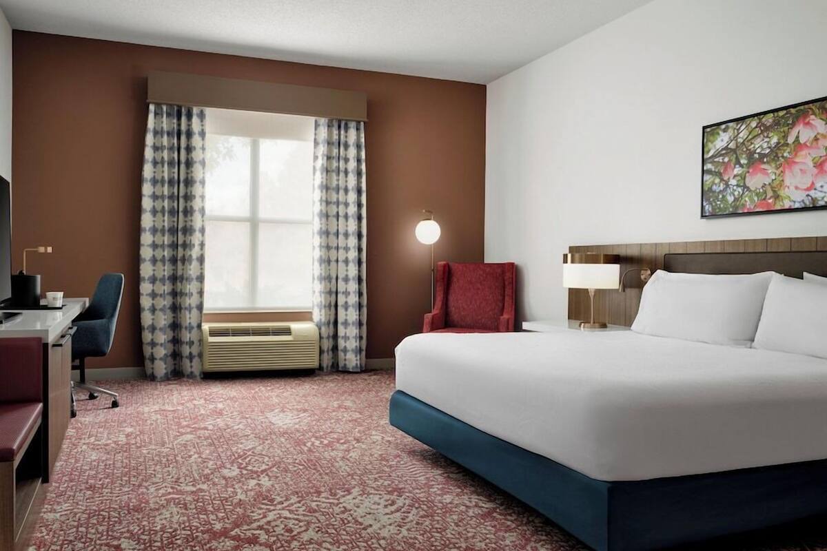 A hotel room with a large bed, a desk, a chair, a lamp, and a window with curtains.
