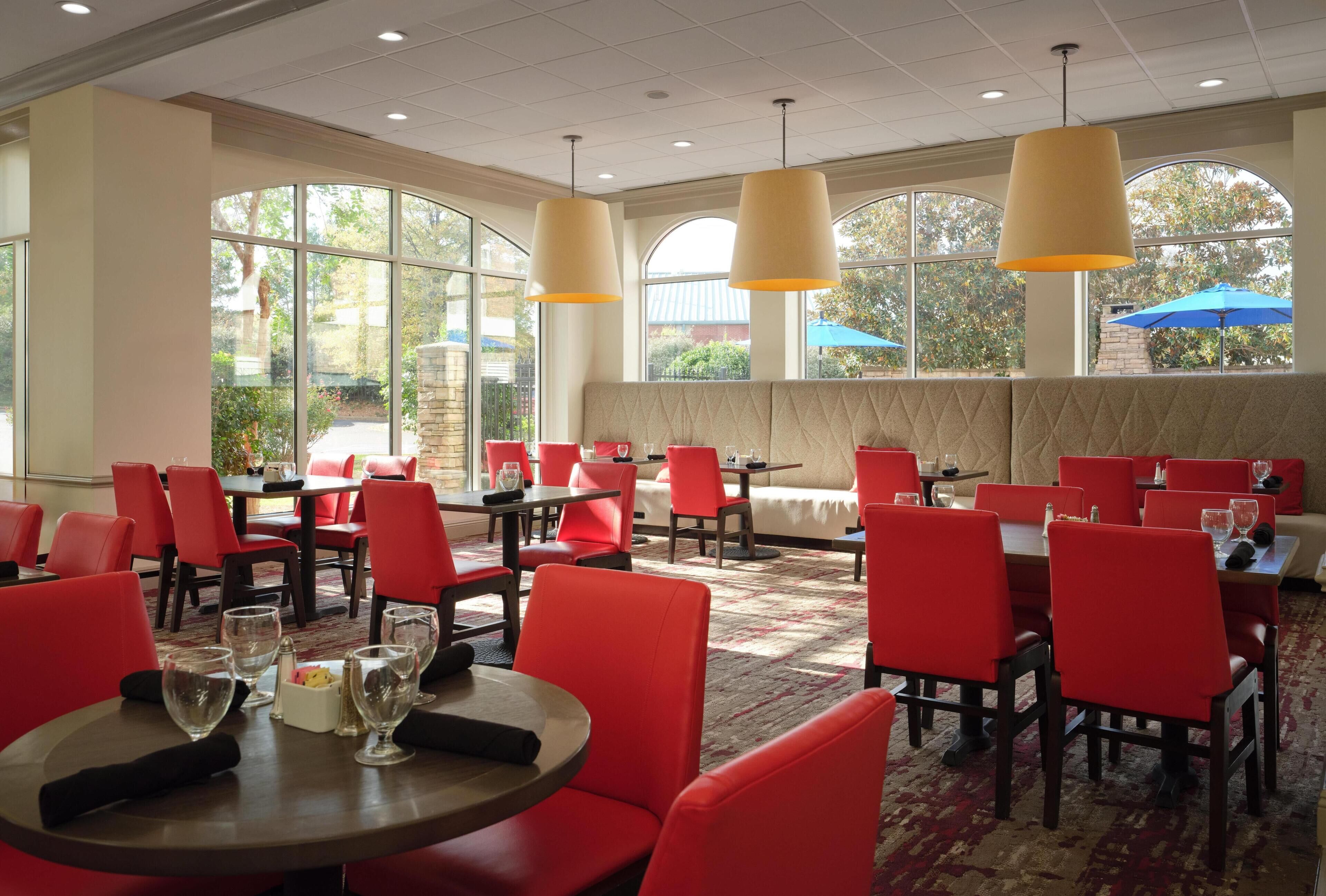 A dining area with red chairs and tables, large windows, and hanging pendant lights.
