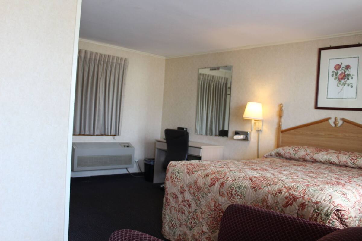 A hotel room with a bed, a desk, a chair, a lamp, a mirror, and a window with curtains.