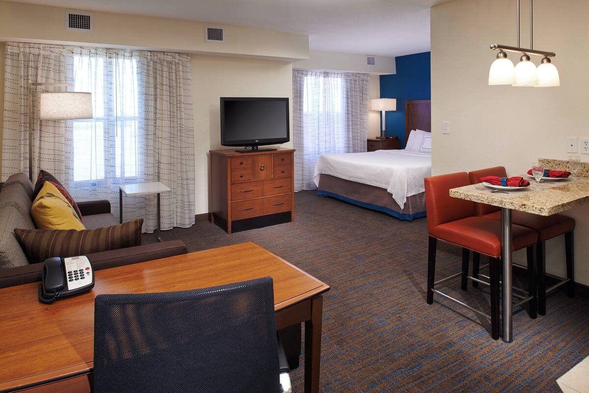 A hotel room with a bed, a TV, a desk with a chair, a small table, and a dining area with chairs.