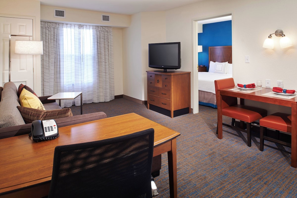 A hotel room with a sofa, a wooden table with chairs, a television on a wooden cabinet, a telephone, and a dining area with a table set for two.