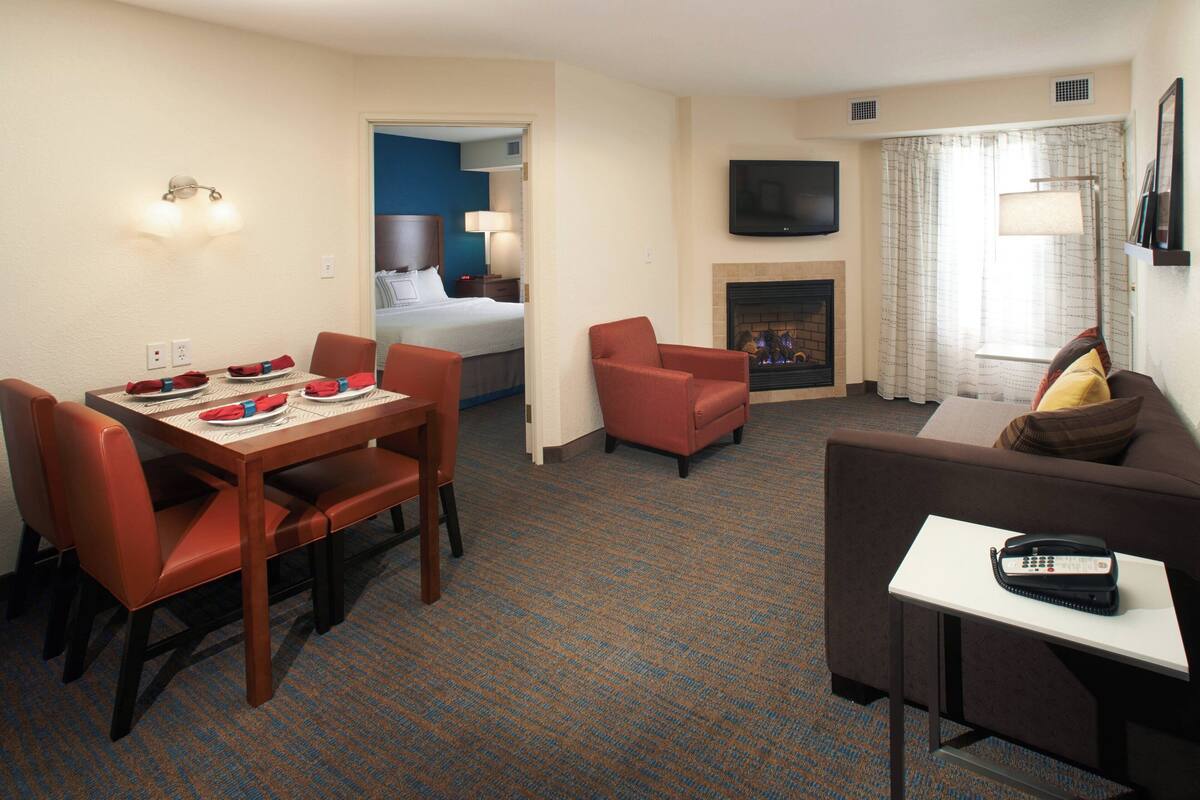 A hotel room with a dining area, a living room with a sofa and armchair, a fireplace, and a TV mounted on the wall.