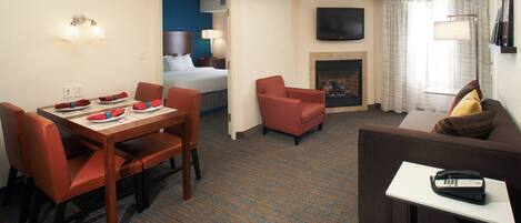 A hotel room with a dining area, a living room with a sofa and armchair, a fireplace, and a TV mounted on the wall.