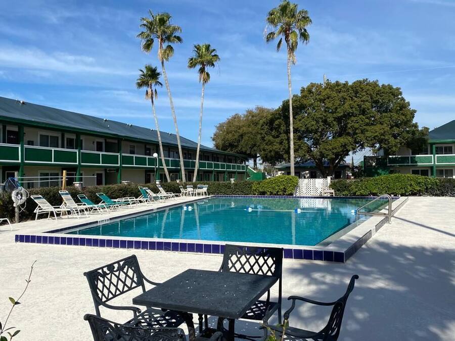 A swimming pool with lounge chairs and a table, surrounded by palm trees and a building with a green roof.