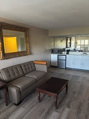 A living room with a sofa, a coffee table, and a kitchen area with cabinets and appliances. - Inn On The Green (Tavares)