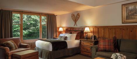 A cozy hotel room with a large bed, a wooden headboard, a desk with a lamp, a sofa, and a window with curtains.