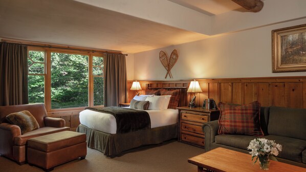 Una habitación de hotel acogedora con una cama grande, un cabecero de madera, un escritorio con lámpara, un sofá y una ventana con cortinas.