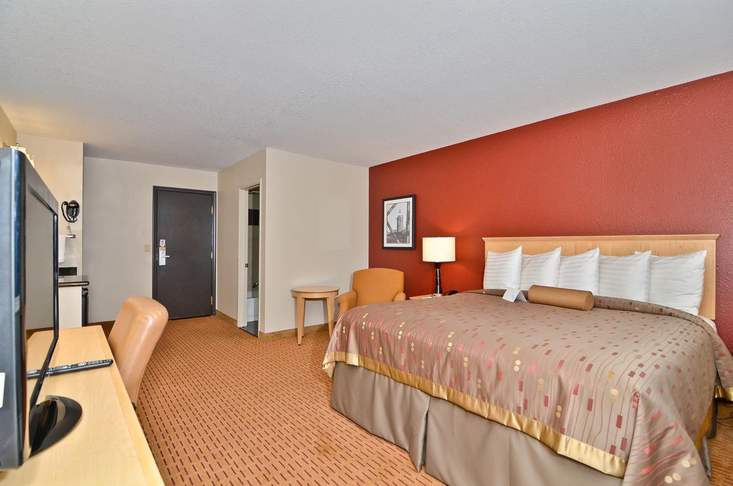 Ein Hotelzimmer mit einem Bett, einem Schreibtisch mit Computer, einem Stuhl, einer Lampe und einer Tür.