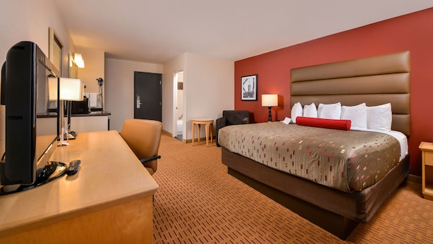 A hotel room with a bed, a desk with a computer and remote controls, a chair, a small table, and a lamp.