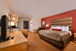 A hotel room with a bed, a desk with a computer and remote controls, a chair, a small table, and a lamp.