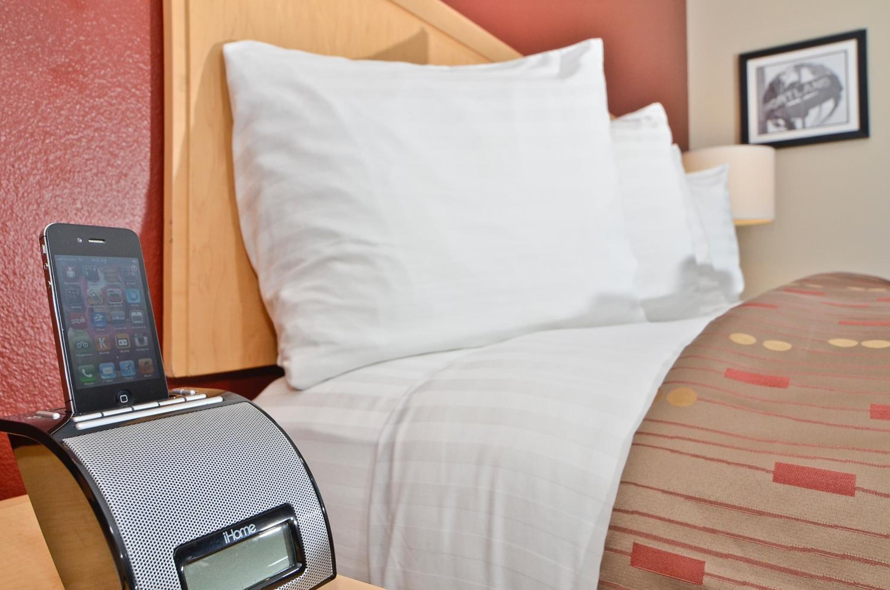 Ein Hotelzimmer mit einem Bett, einem weißen Kopfkissen, einer gemusterten Decke und einem tragbaren Lautsprecher, auf dem ein Smartphone liegt.