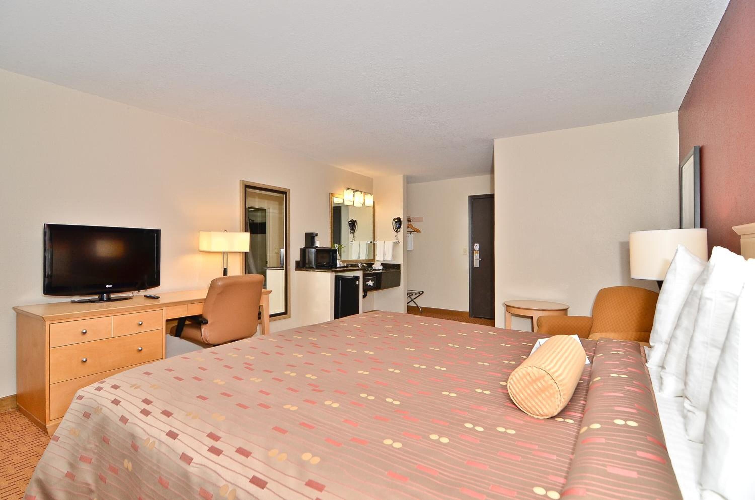 Ein Hotelzimmer mit Bett, Schreibtisch, Fernseher, Spiegel, einer kleinen Küchenzeile und einer Tür.