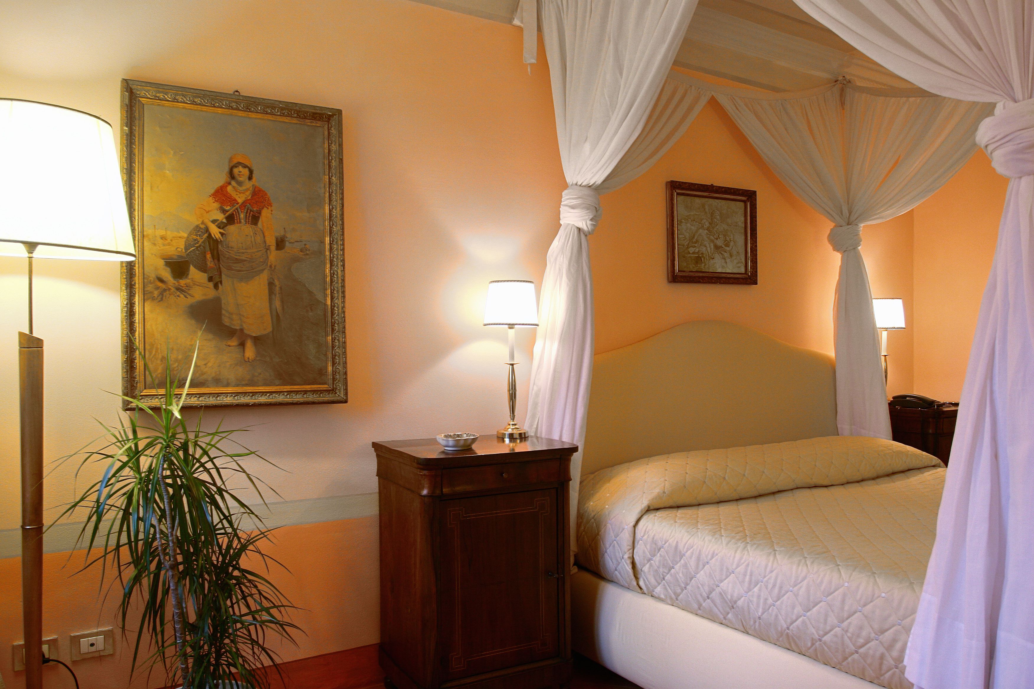 A bedroom with a bed, a nightstand, a lamp, a painting, and a potted plant.