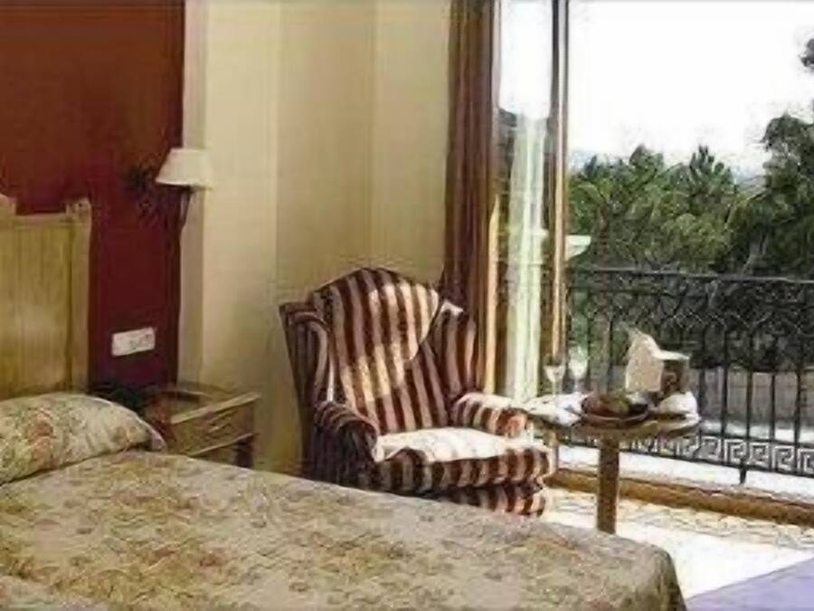 A hotel room with a bed, a chair, a nightstand, and a window with a view of greenery.