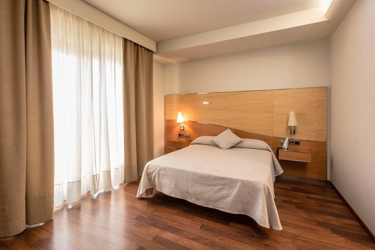 A hotel room with a bed, wooden headboard, bedside lamps, and a window with curtains.