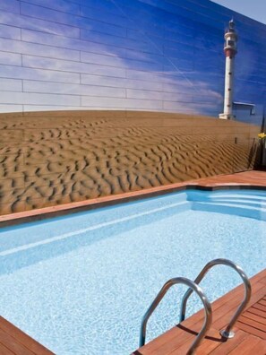 A swimming pool with a wooden deck, a lighthouse in the background, and a sandy area beside the pool.