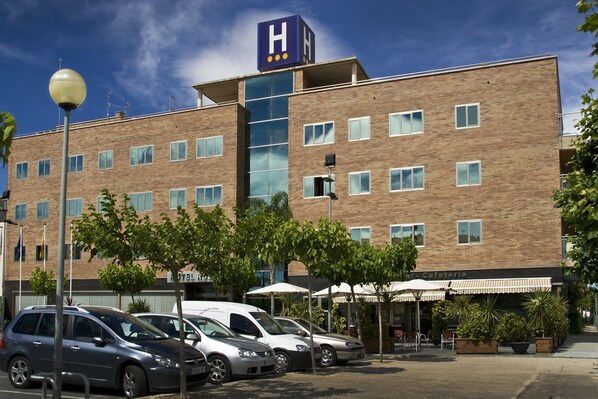 A hotel with a prominent 'H' sign, multiple windows, and a parking area with cars. - Hotel Rull (Deltebre)