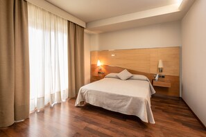 A hotel room with a large bed, wooden headboard, bedside lamps, and a window with curtains.