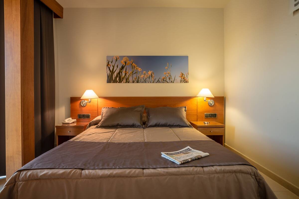 A neatly made bed with a magazine, two bedside lamps, a wooden headboard, and a wall-mounted painting.