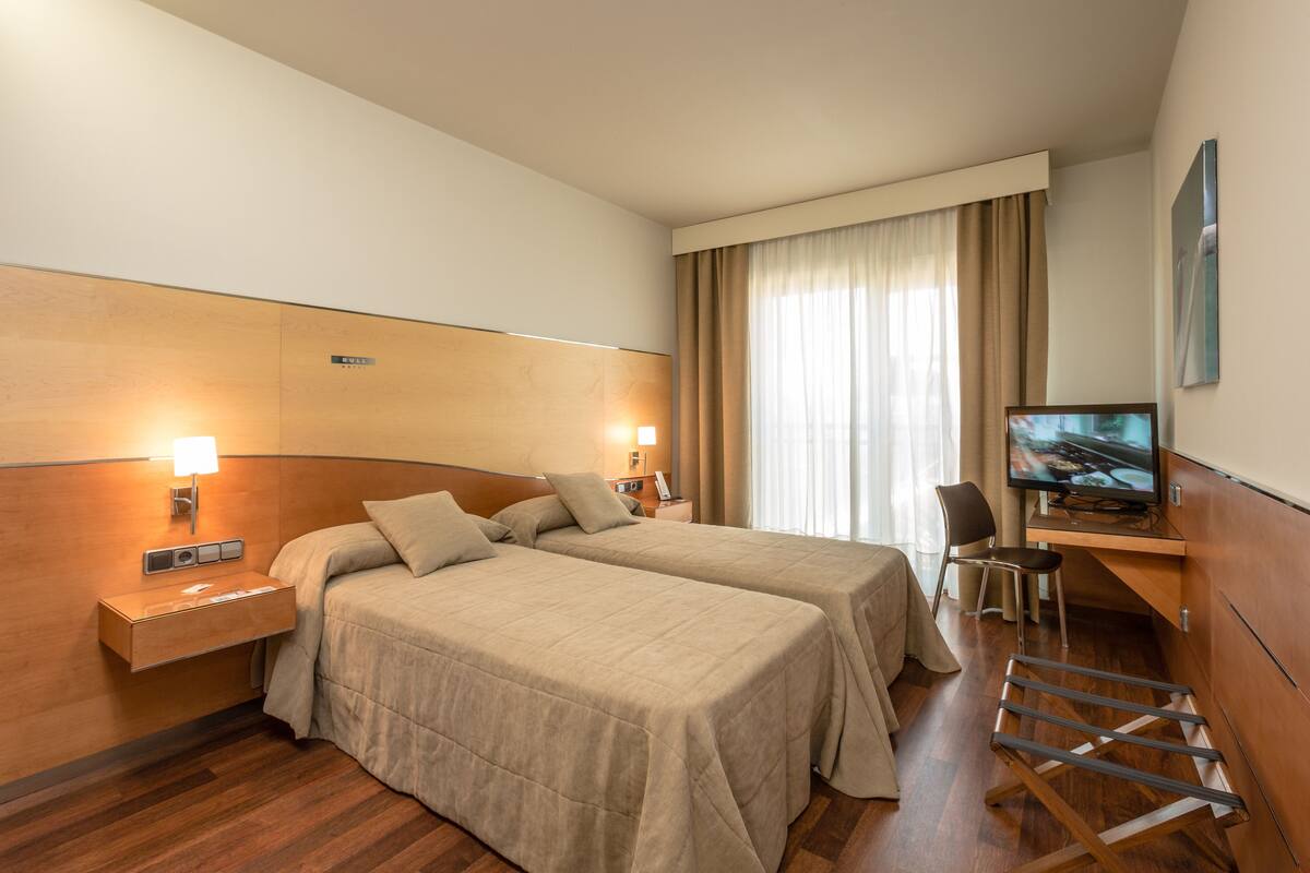 A hotel room with two beds, a desk, a chair, a television, and a window with curtains.