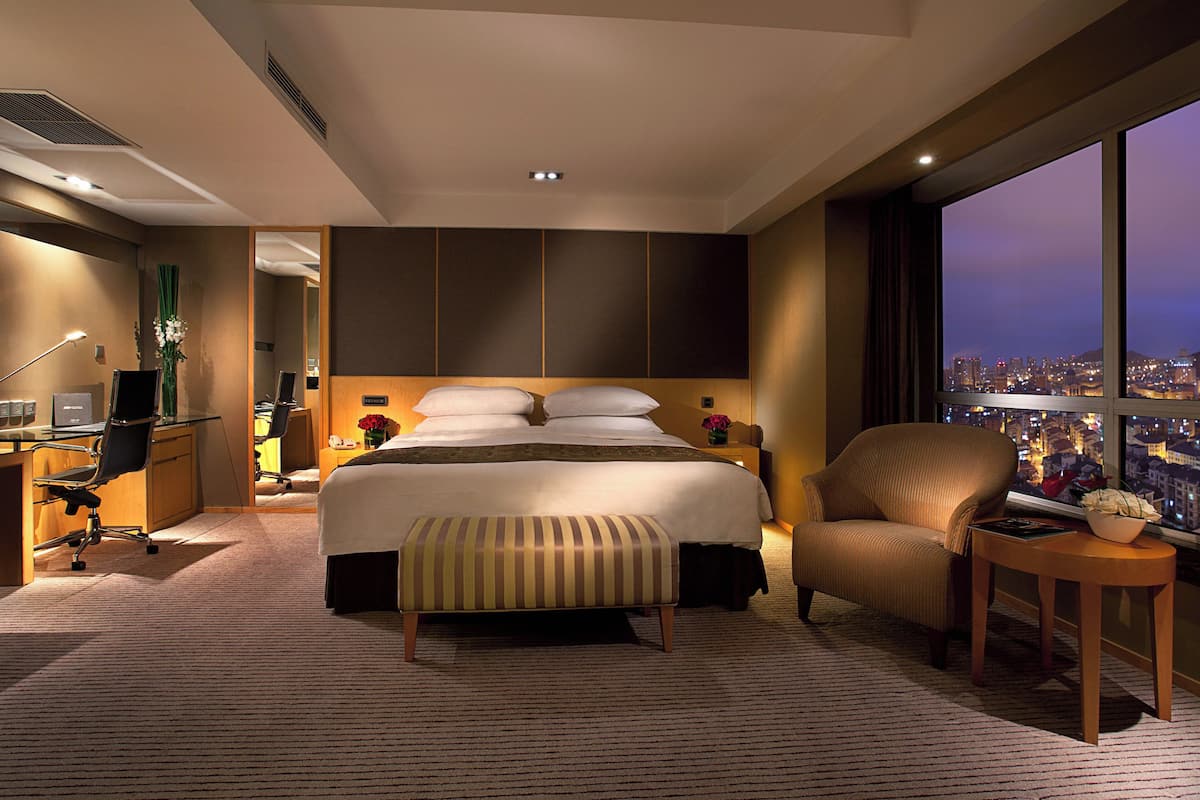 Ein modernes Hotelzimmer mit einem großen Bett, einem Schreibtisch, einem Stuhl und Blick auf die beleuchtete Stadt.