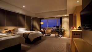 Habitación de hotel con dos camas, un escritorio, una silla, un televisor y vista a la ciudad de noche.