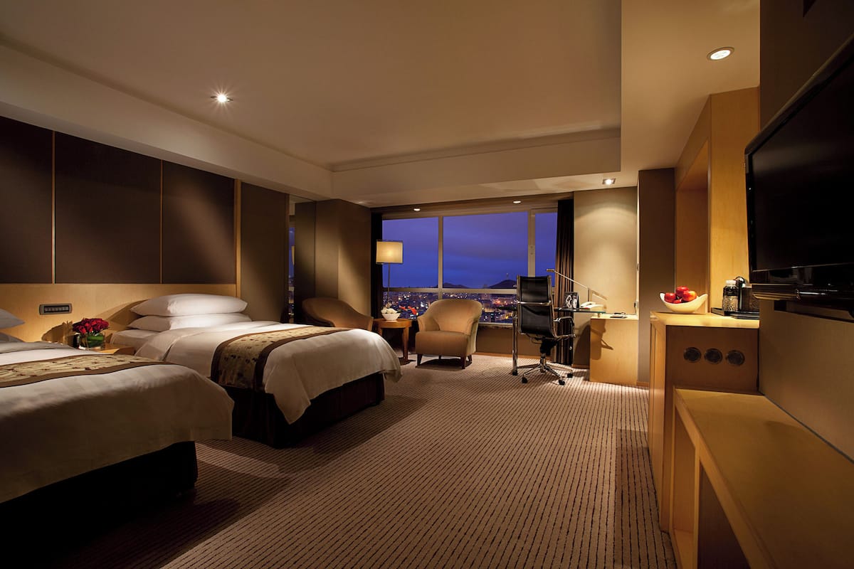 Ein Hotelzimmer mit zwei Betten, einem Schreibtisch, einem Sessel, einem Fernseher und Blick auf die nächtliche Stadt.