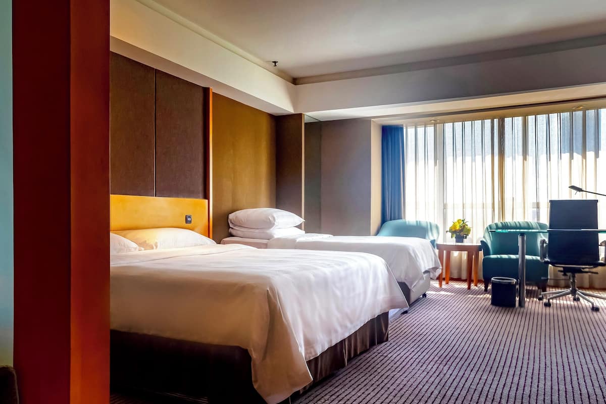 Ein Hotelzimmer mit zwei Betten, einem Schreibtisch mit Stuhl, einem kleinen Tisch mit Pflanze und einem großen Fenster mit Vorhängen.