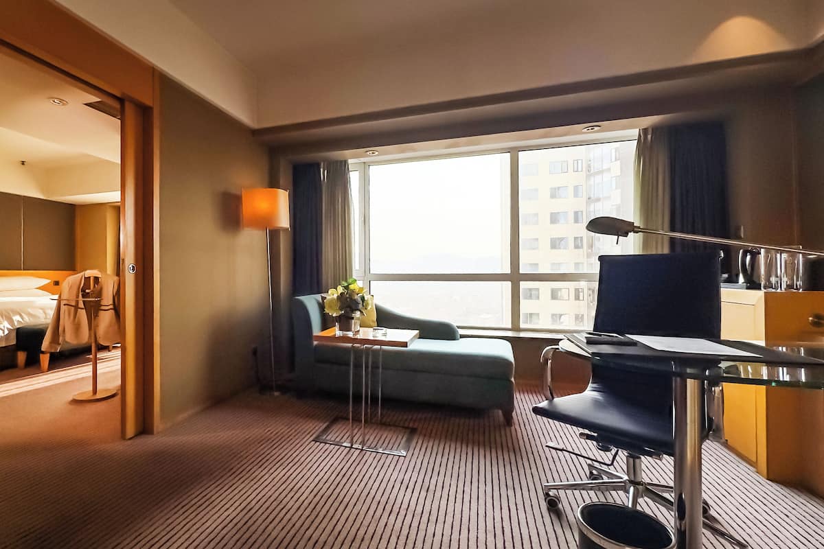 Ein Hotelzimmer mit einem Bett, einem Schreibtisch mit Stuhl, einem Sofa, einem Fenster mit Blick auf Gebäude und einer Tür zu einem anderen Raum.