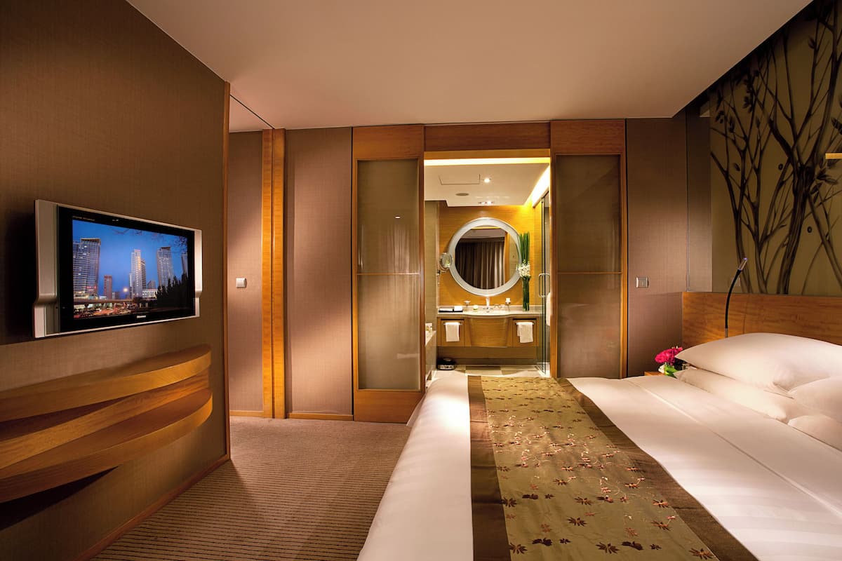 Ein modernes Hotelzimmer mit einem Flachbildfernseher, einem großen Bett mit floralem Überwurf, einem Holz-Kopfteil und einem großen Spiegel über dem Schminktisch.