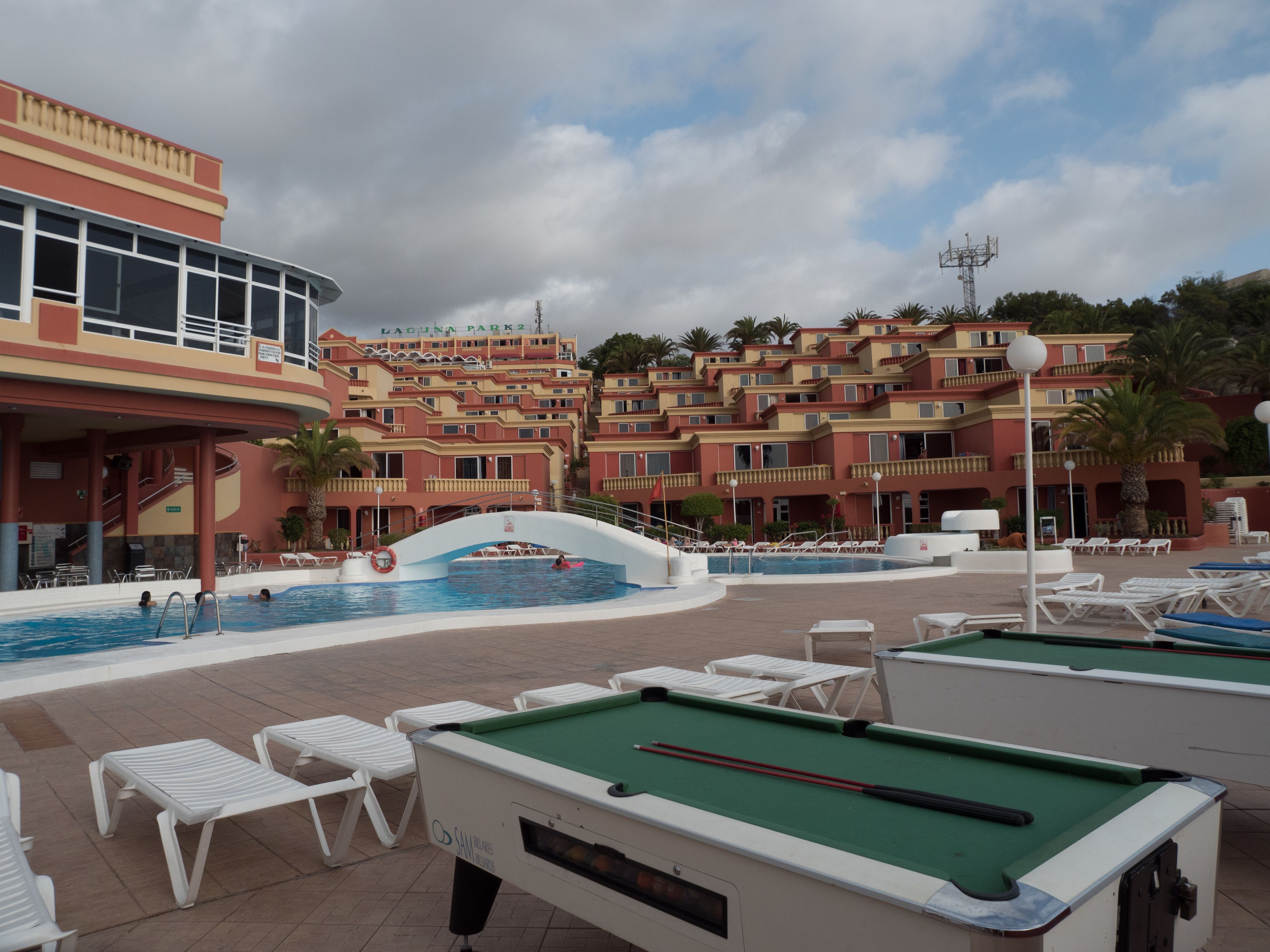 Un área de piscina con sillas de descanso, mesa de billar y un edificio con balcones.