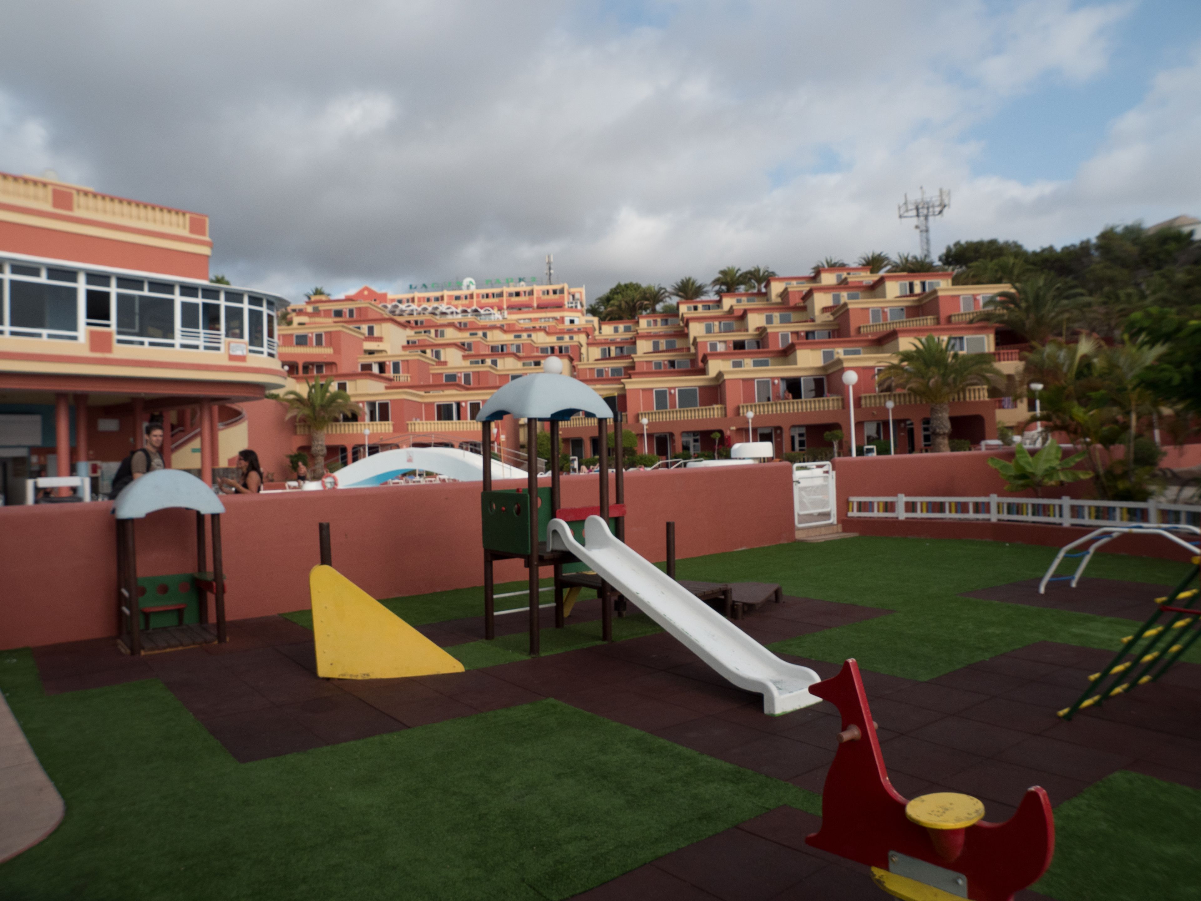 A playground with slides and climbing structures, surrounded by a red wall and green artificial grass.