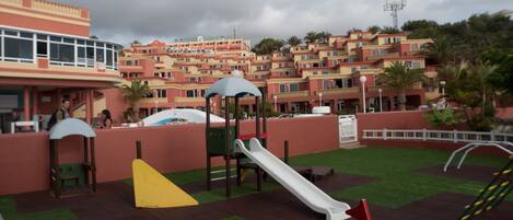 A playground with slides and climbing structures, surrounded by a red wall and green artificial grass.