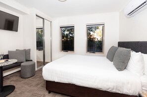 Ein modernes Schlafzimmer mit einem großen Bett, einem Schreibtisch mit Stuhl und einem kleinen Hocker. Zwei Fenster sorgen für natürliches Licht und einen Ausblick ins Freie.