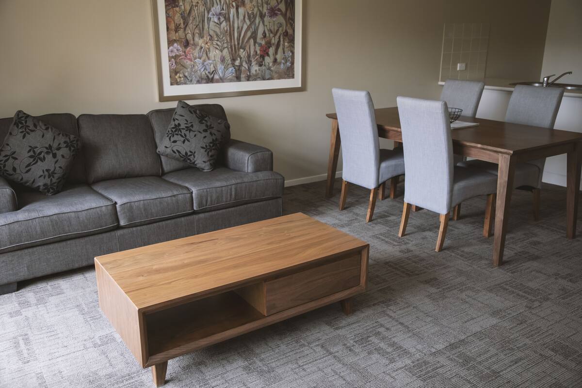Een woonkamer met een grijze bank, een houten salontafel en een eethoek met een houten tafel en stoelen.