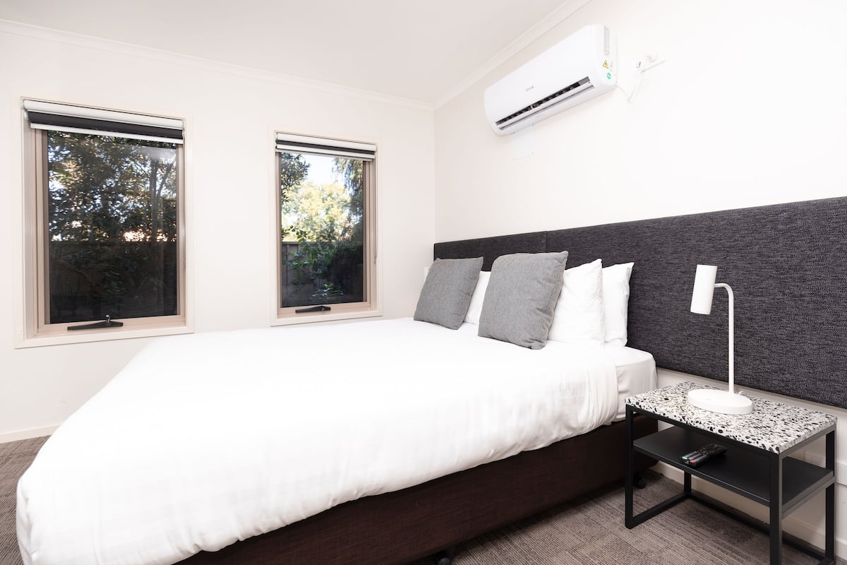 Een moderne slaapkamer met een groot bed, twee ramen, een nachtkastje met een lamp en een airconditioning.
