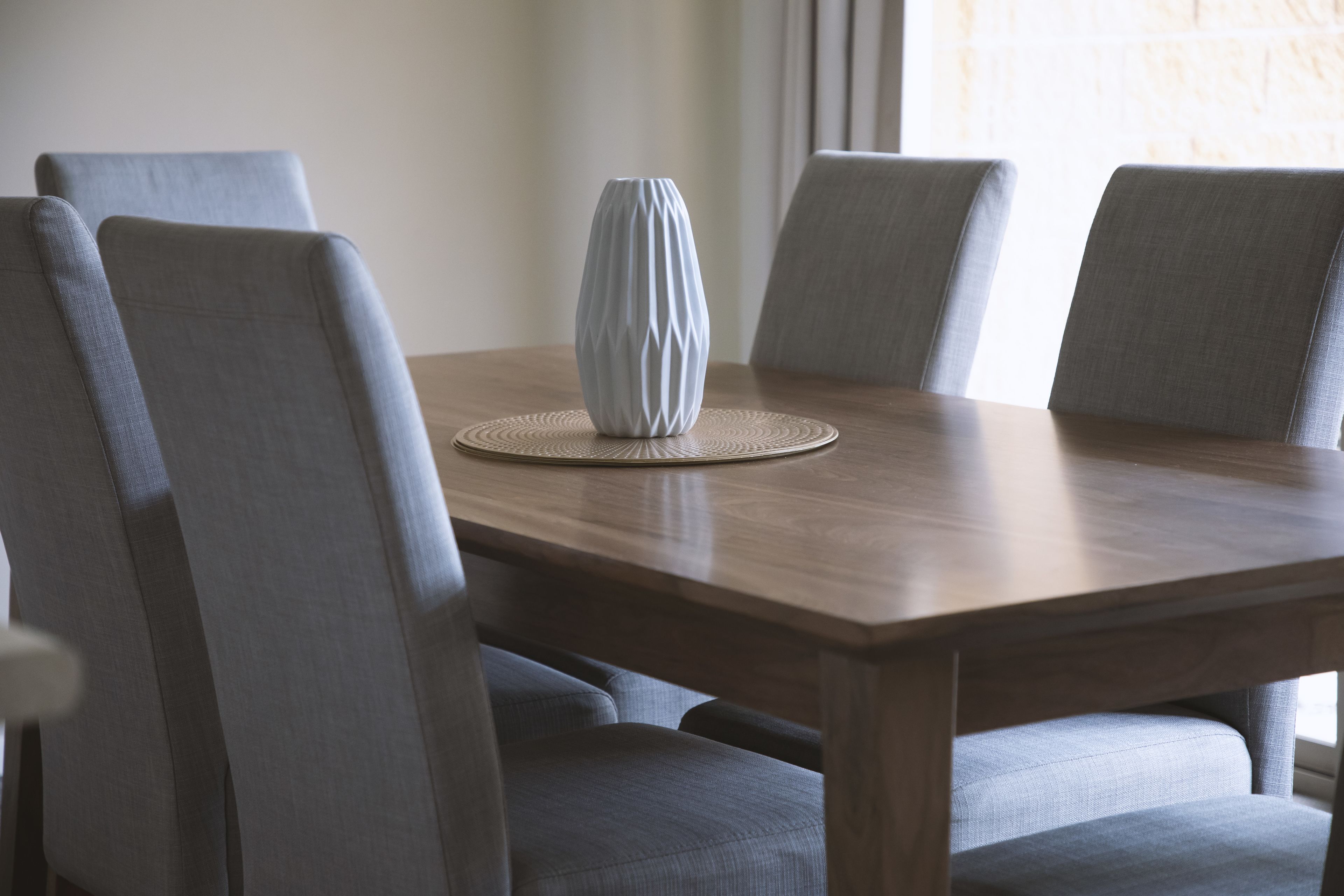 A dining table with four grey upholstered chairs, a white vase, and a woven placemat.