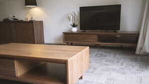 Un salon moderne avec une table basse en bois, un téléviseur posé sur un meuble en bois et un petit vase blanc contenant une orchidée.