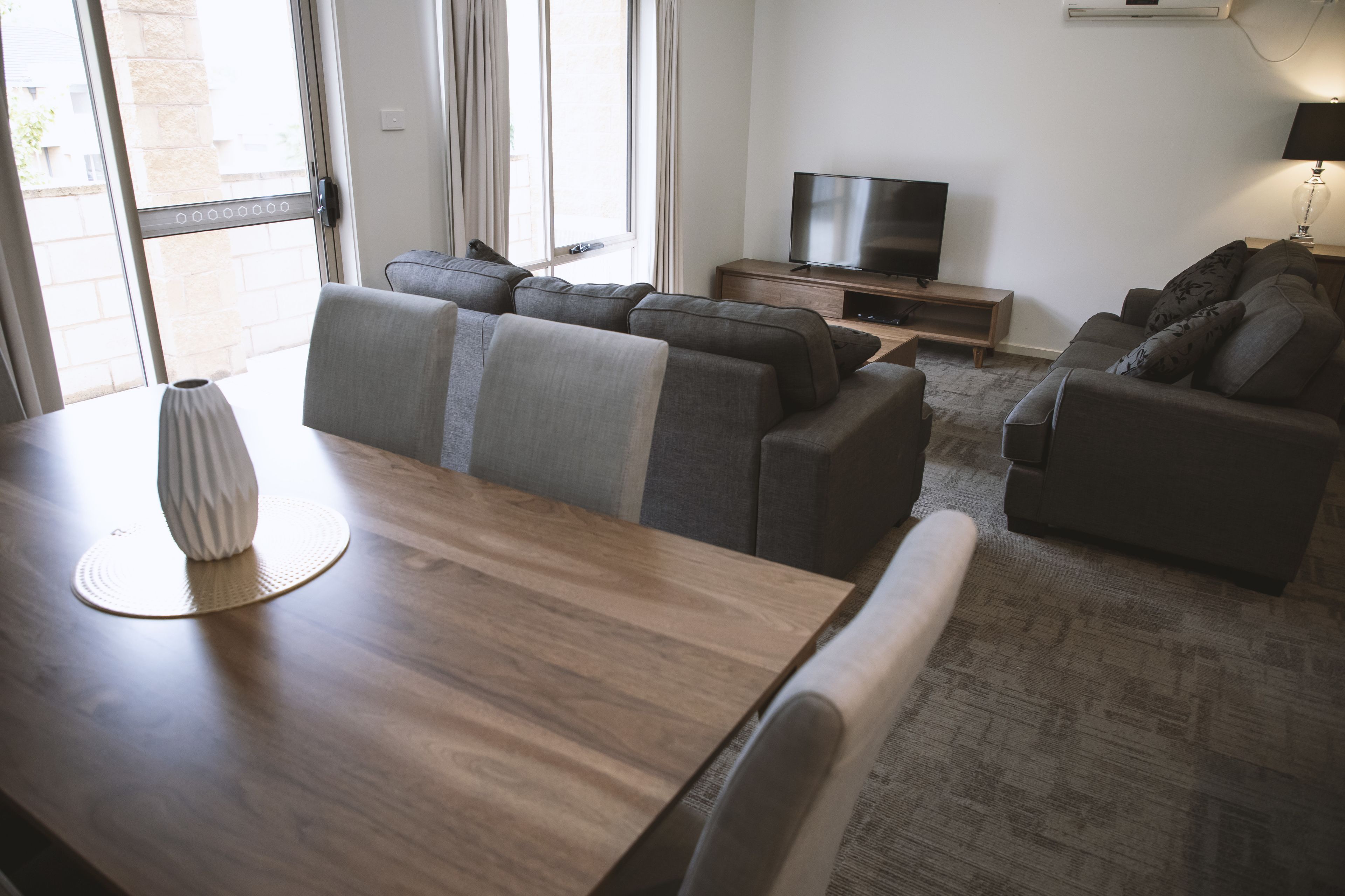 Un salon moderne avec une table à manger en bois, des canapés gris, un téléviseur à écran plat et une lampe.